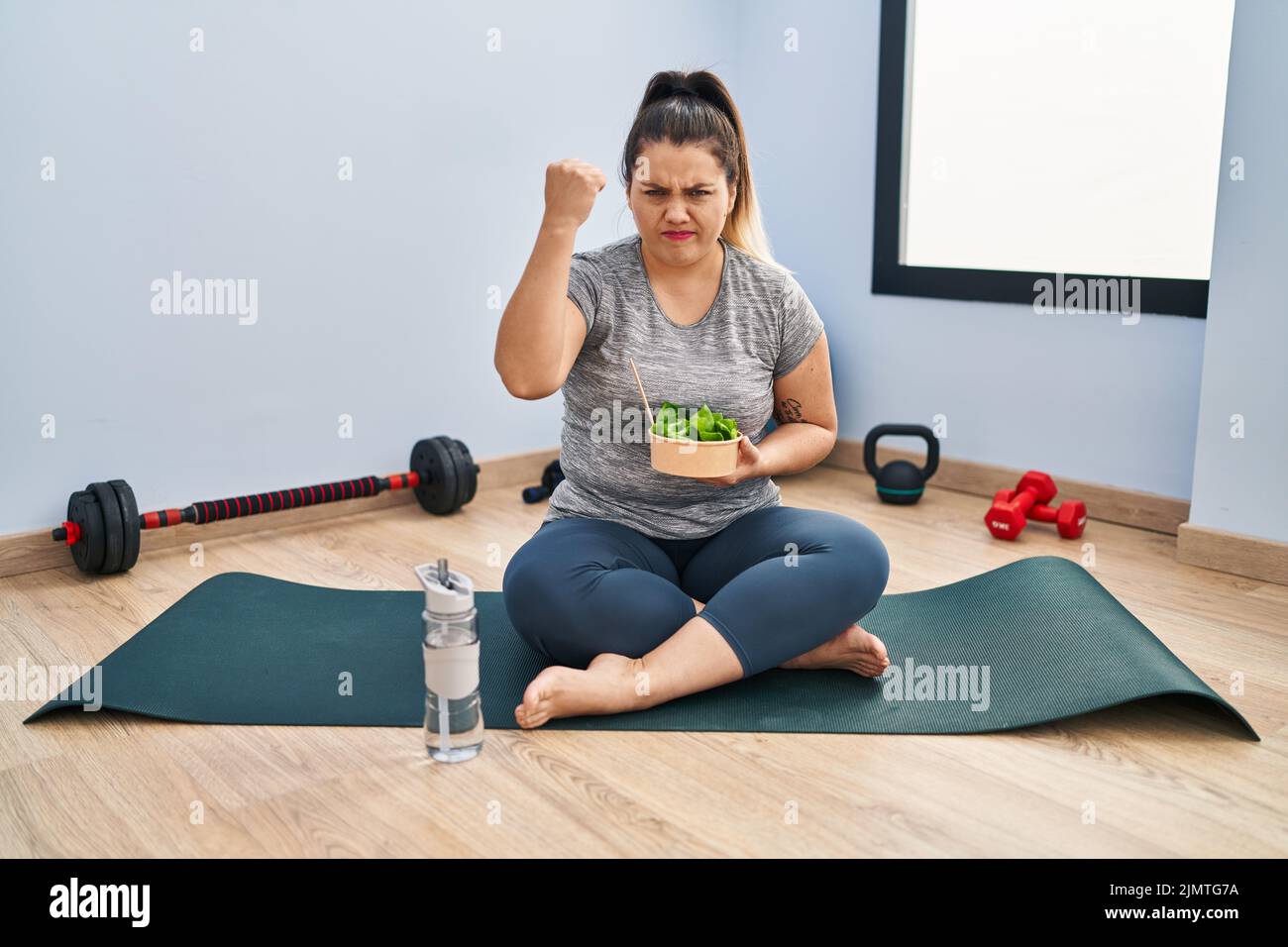 Young hispanic woman sitting on yoga mat eating healthy salad annoyed ...