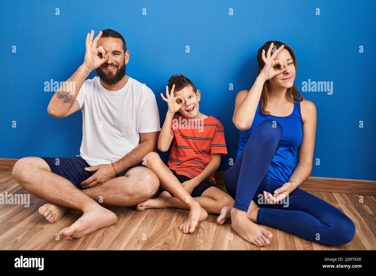 Family of three sitting on the floor at home doing ok gesture with hand smiling, eye looking through fingers with happy face. Stock Photo