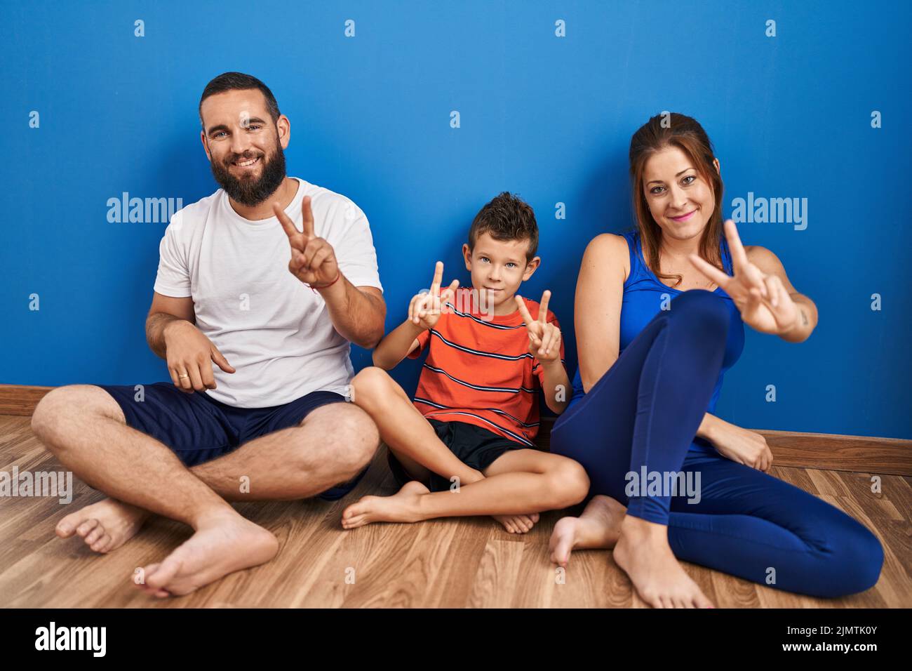 Family of three sitting on the floor at home smiling looking to the camera showing fingers doing victory sign. number two. Stock Photo