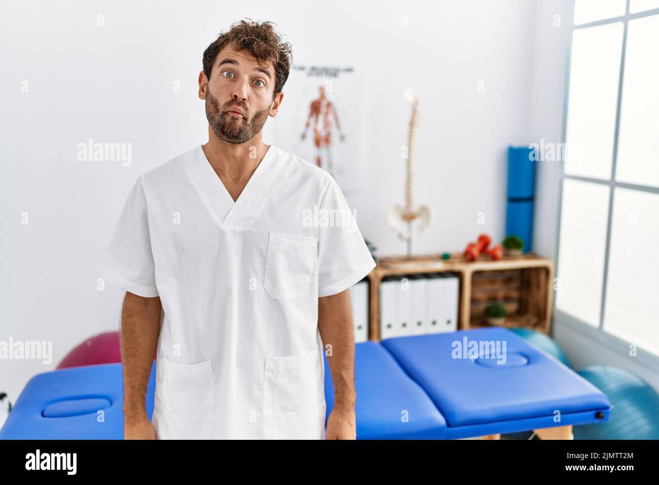 Young handsome physiotherapist man working at pain recovery clinic making fish face with lips, crazy and comical gesture. funny expression. Stock Photo