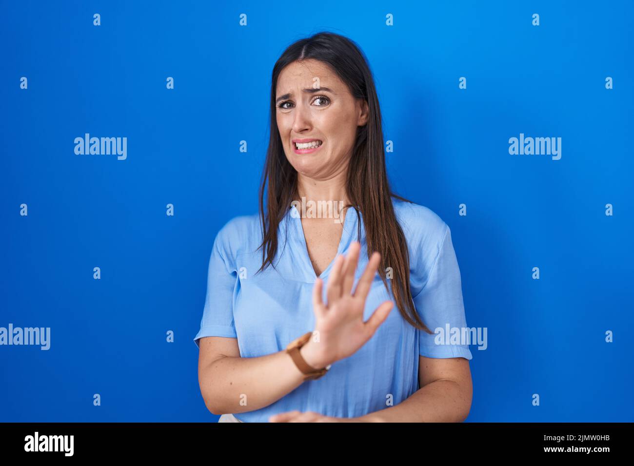 Young brunette woman standing over blue background disgusted expression ...