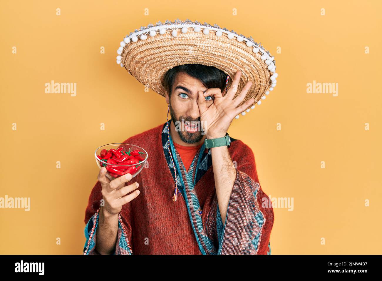 Young hispanic man wearing mexican hat holding chili smiling happy doing ok sign with hand on eye looking through fingers Stock Photo