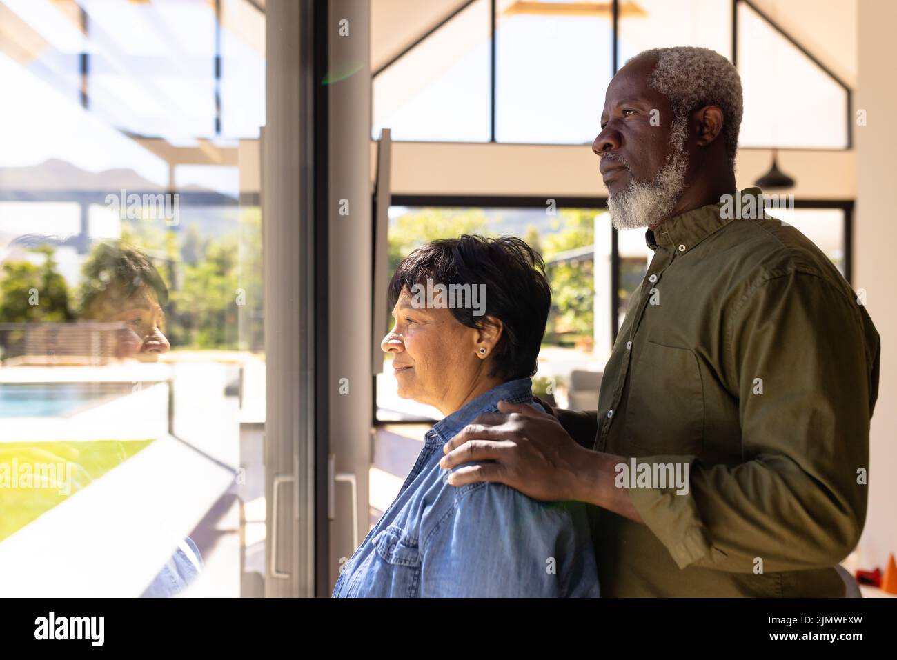 Side view of multiracial senior couple looking through window while standing in retirement home Stock Photo