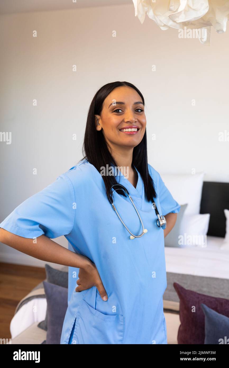 Portrait of smiling biracial female physiotherapist with arms akimbo standing against wall at home Stock Photo