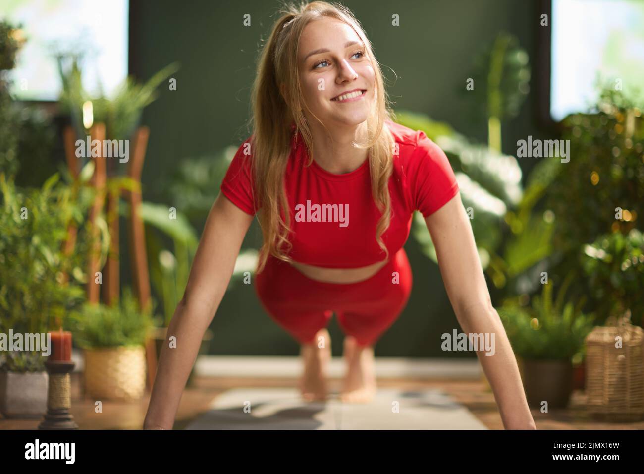 smiling young woman in red fitness clothes at modern green home training. Stock Photo