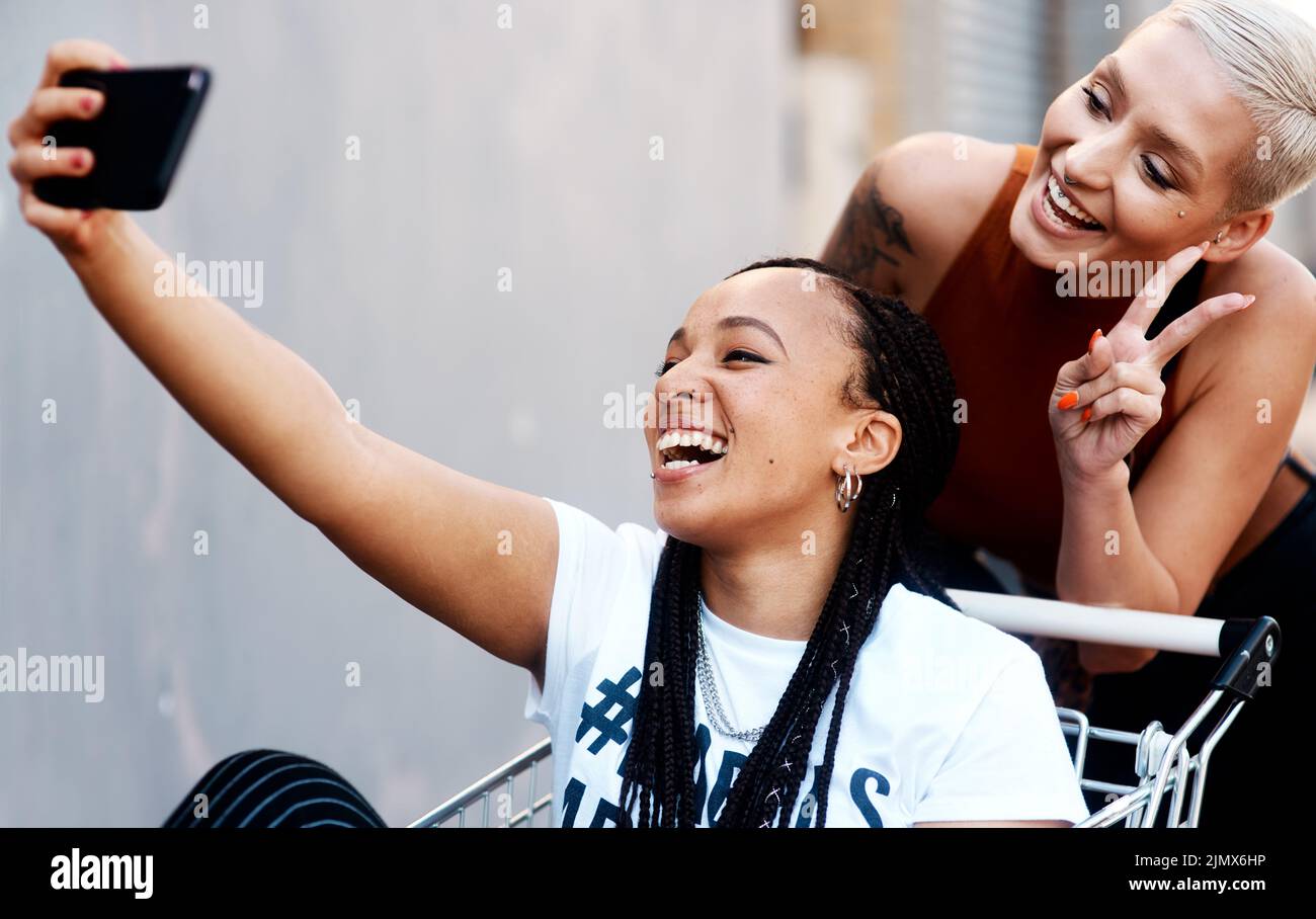 Peace out to you, from my best friend and I. two cheerful young girlfriends taking a selfie while playing around with a shopping cart outdoors. Stock Photo