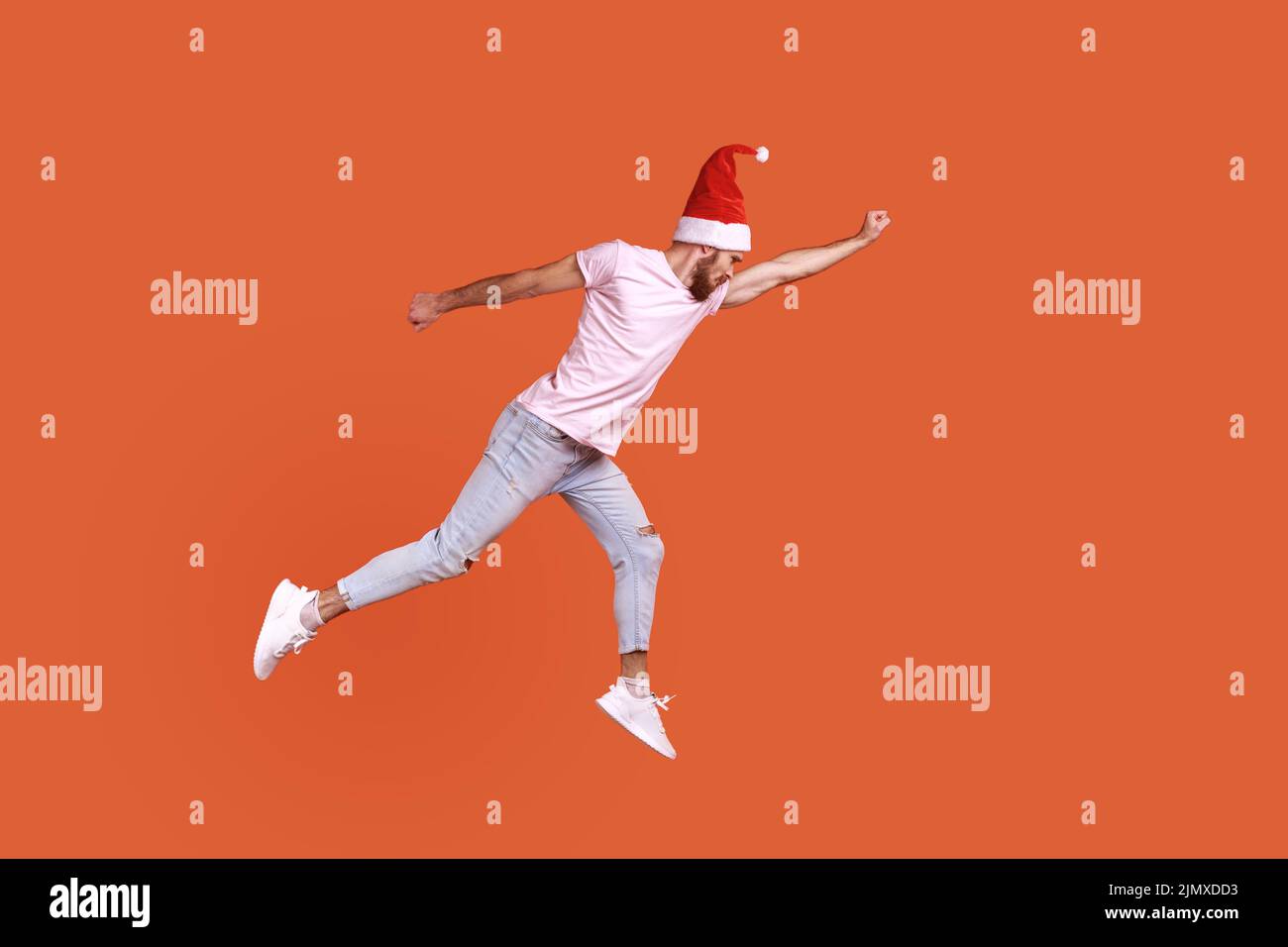 Full length of man in santa hat flying up in air as superhero with raised hand, feeling superpower, being hurry to celebrate New year, wearing T-shirt. Indoor studio shot isolated on orange background Stock Photo