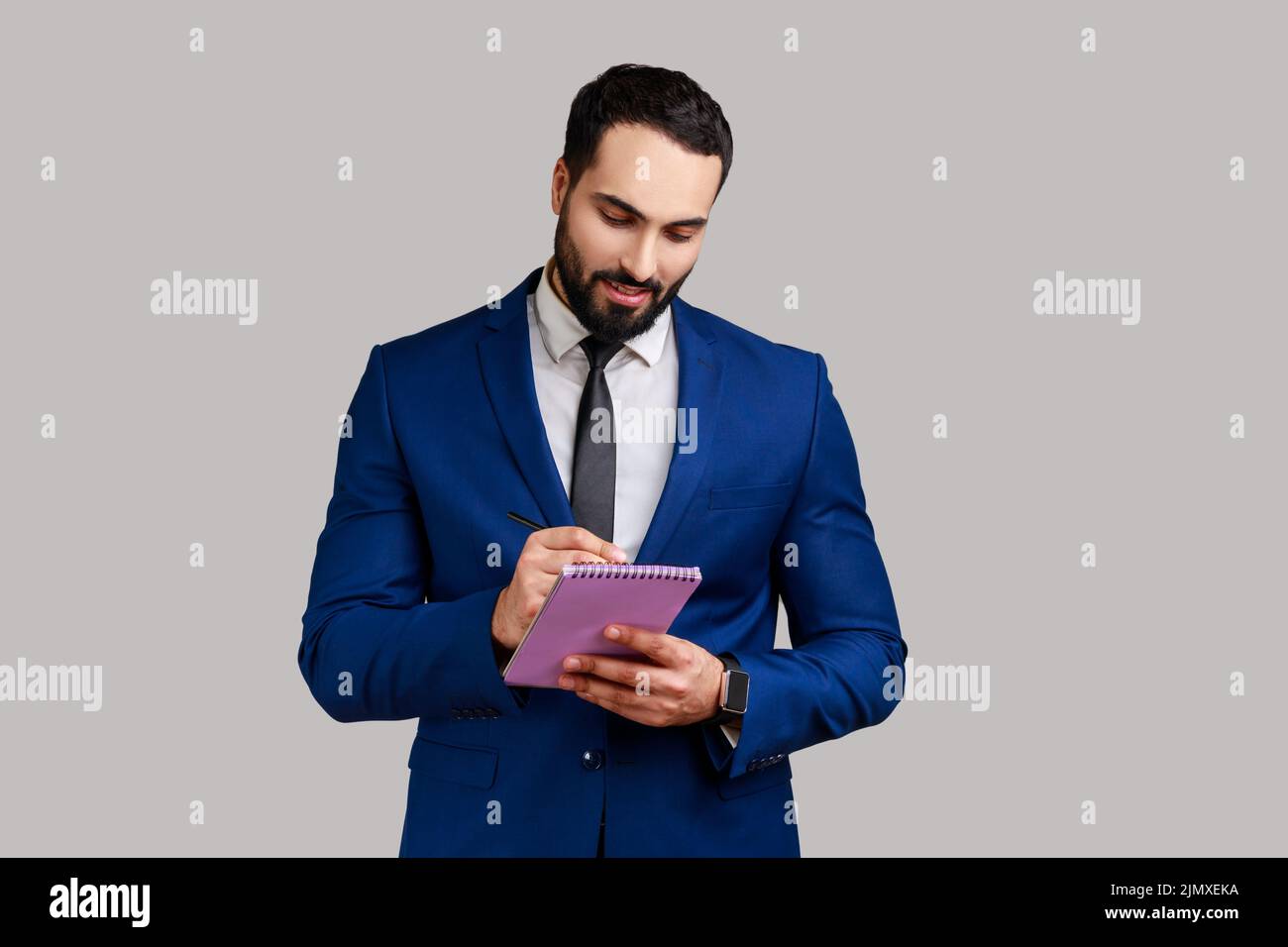 Portrait of positive bearded businessman writing down in paper notebook, making to do list, having good mood, wearing official style suit. Indoor studio shot isolated on gray background. Stock Photo