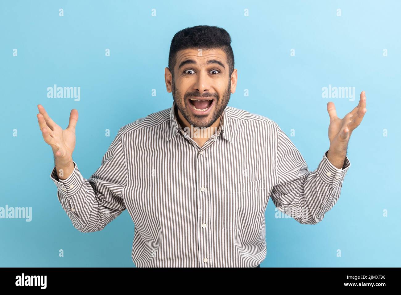 Irritated annoyed businessman with beard has problems, gesticulating with hands, crisis and negativity, misunderstanding, wearing striped shirt. Indoor studio shot isolated on blue background. Stock Photo