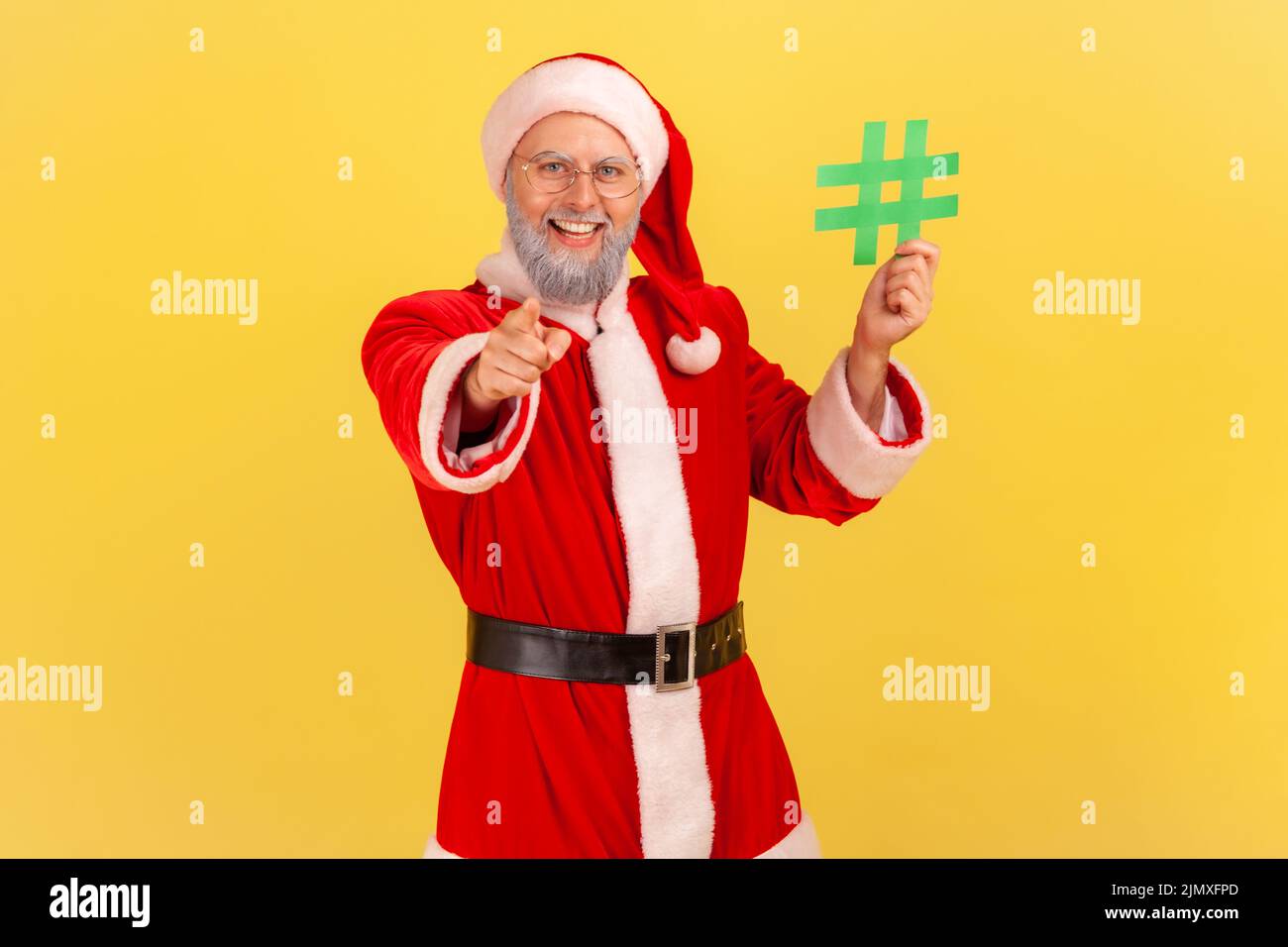 Positive elderly man with gray beard wearing santa claus costume standing with green hashtag in hands, pointing to camera with toothy smile. Indoor studio shot isolated on yellow background. Stock Photo