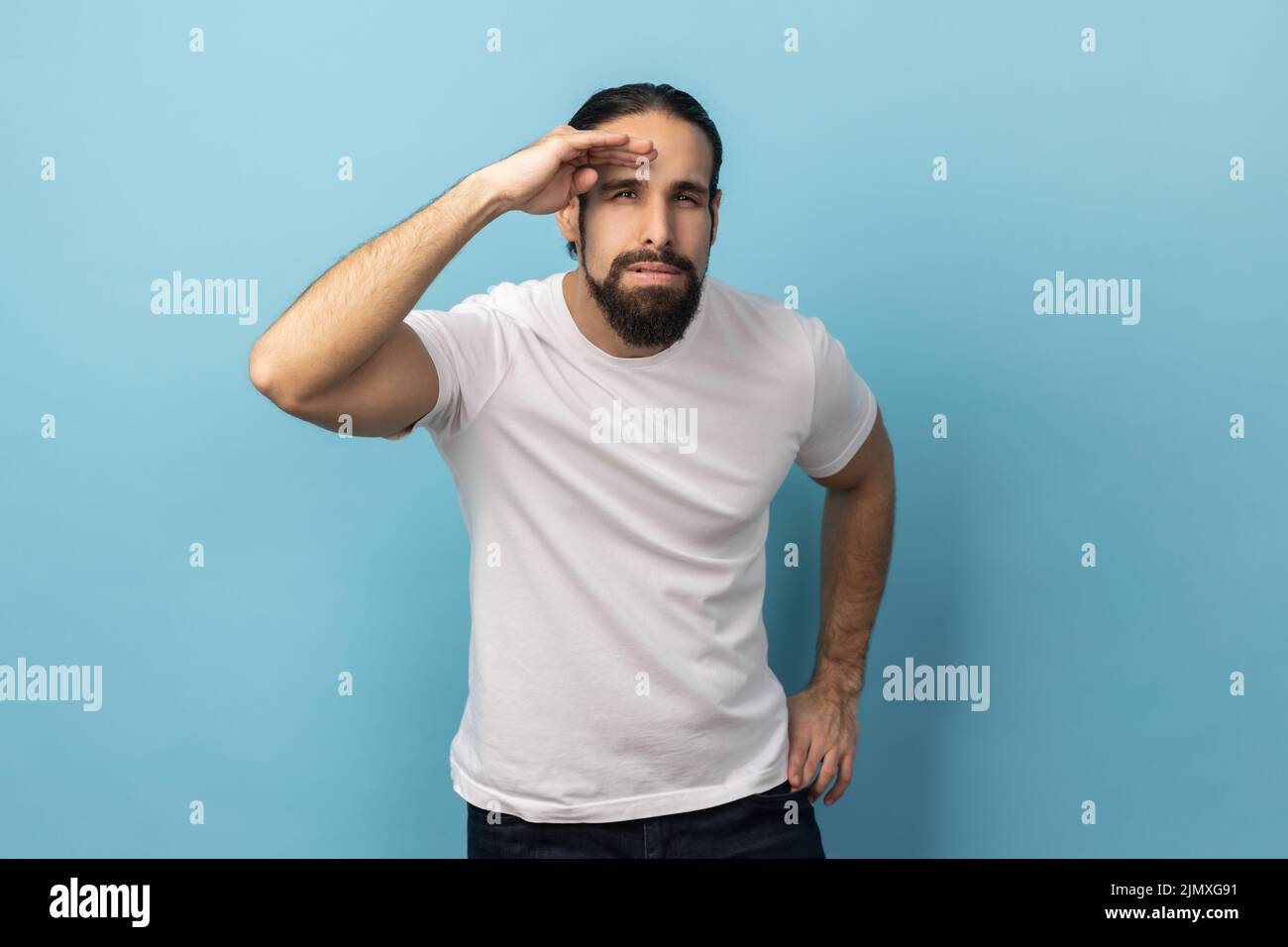 Portrait of curious man with beard wearing T-shirt keeping palm over head and looking attentively far away, peering with expectation at long distance. Indoor studio shot isolated on blue background. Stock Photo