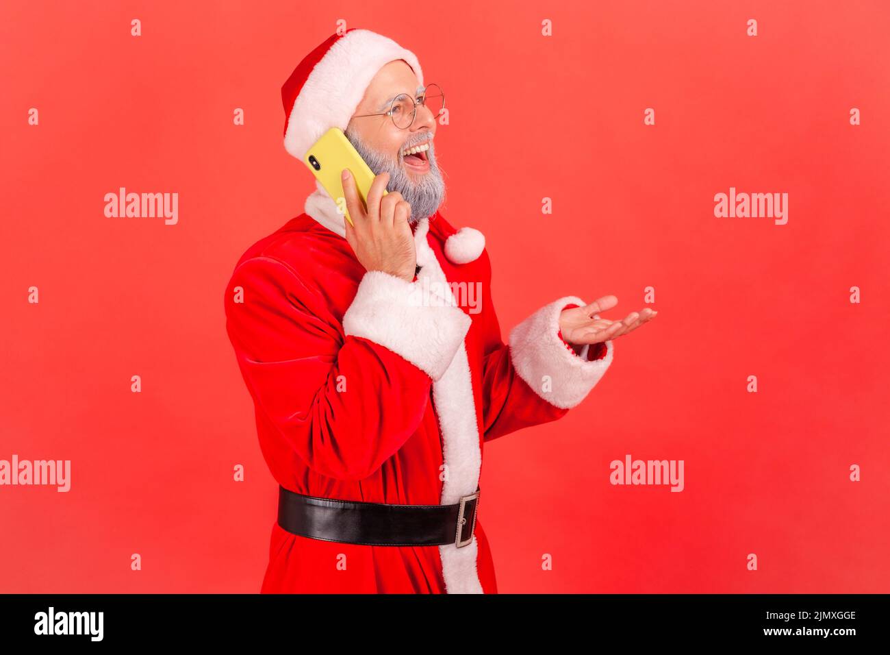 Happy smiling elderly man with gray beard wearing santa claus costume talking phone, speaking with positive expression, pleasant news. Indoor studio shot isolated on red background. Stock Photo