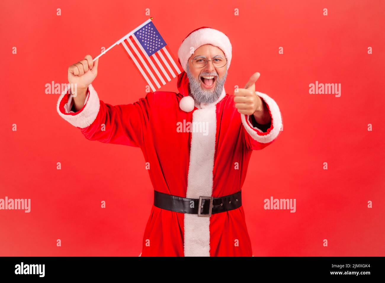 Smiling positive elderly man with gray beard wearing santa claus costume holding USA flag, showing thumb up to camera, having patriotic mood. Indoor studio shot isolated on red background. Stock Photo