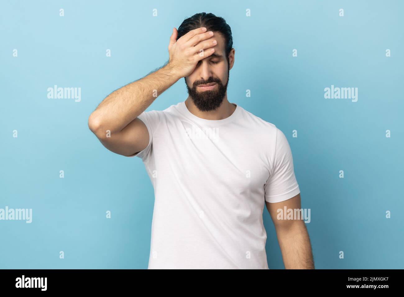 Portrait of unlucky man with beard wearing white T-shirt standing with facepalm gesture, feeling regret and sorrow, blaming herself for mistake. Indoor studio shot isolated on blue background. Stock Photo