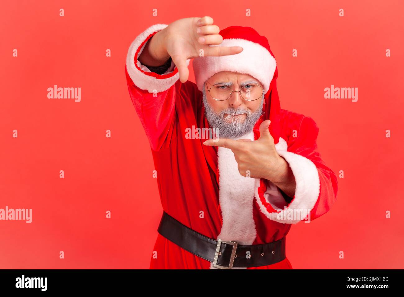 Elderly man in santa claus costume gesturing picture frame with hands, looking through fingers and focusing on interesting moment, taking photo. Indoor studio shot isolated on red background. Stock Photo