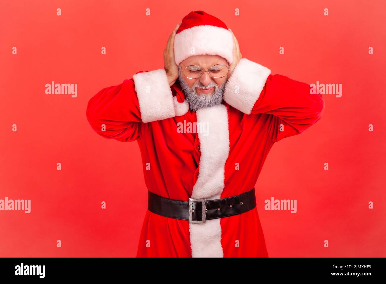 Don't want here. Elderly man with gray beard wearing santa claus costume standing covering ears with palms, loud noise, frowning face. Indoor studio shot isolated on red background. Stock Photo