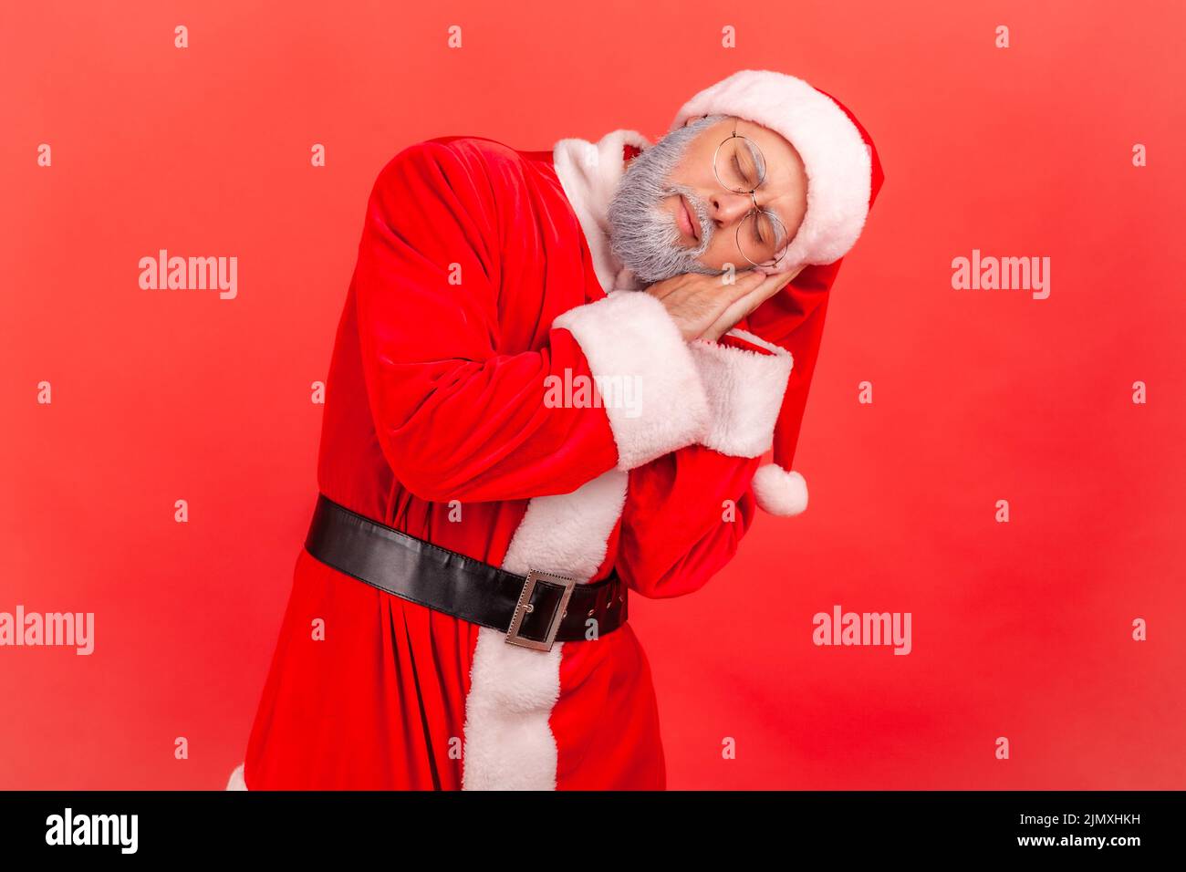 Elderly man wearing santa claus costume sleeping laying down on her palms and smiling pleased, having comfortable nap and resting, dozing off. Indoor studio shot isolated on red background. Stock Photo