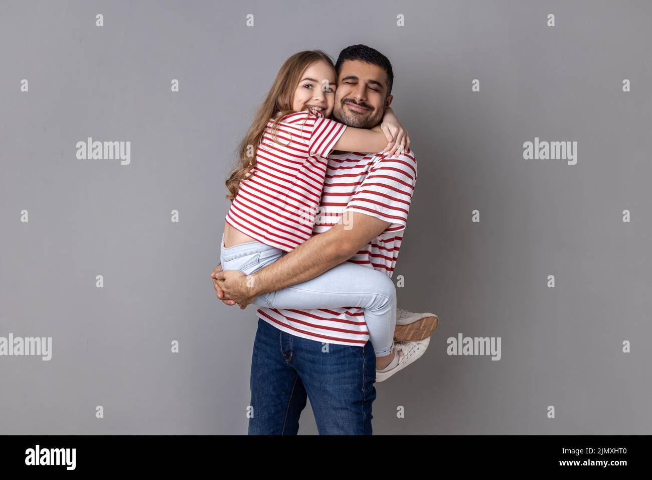 Portrait of father and daughter in striped T-shirts holding his child, feeling love and deep devotion, warm relationships, love care, fathers day. Indoor studio shot isolated on gray background. Stock Photo