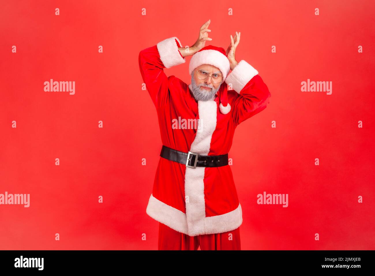 I am deer. Elderly man with gray beard wearing santa claus costume showing deer antler horns over head, looking with comical humorous expression. Indoor studio shot isolated on red background. Stock Photo