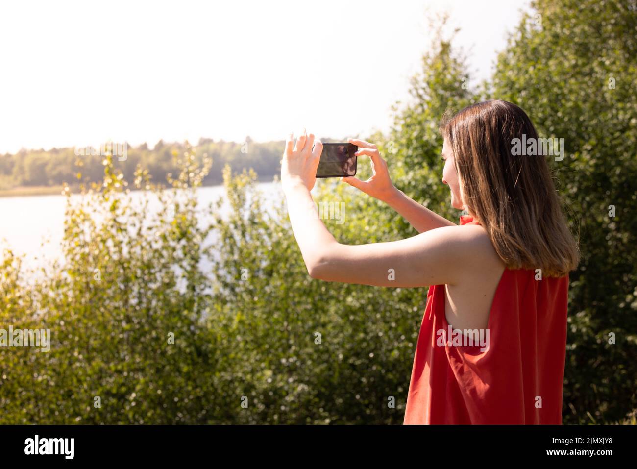 The pretty young caucasian woman in red skirt is taking taking photo by mobile phone in the green forest lakeside at the outside Stock Photo