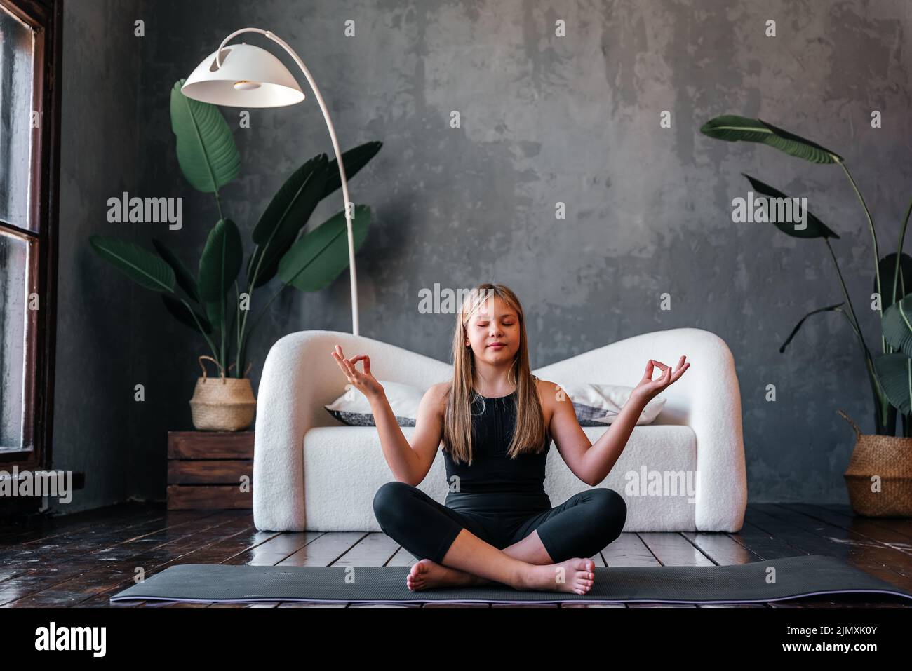 Young attractive girl practicing yoga, sitting in Half Lotus exercise, Ardha Padmasana pose, exercising, meditation session, home interior. High quali Stock Photo