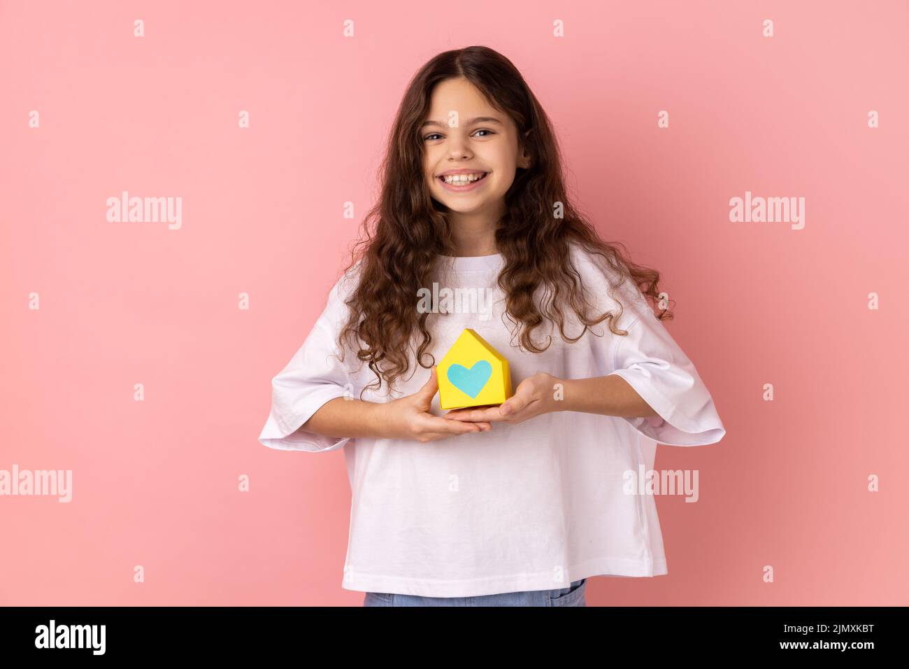 Portrait of smiling charming little girl wearing white T-shirt holding out paper small house, planning new home to buy, real estate. Indoor studio shot isolated on pink background. Stock Photo