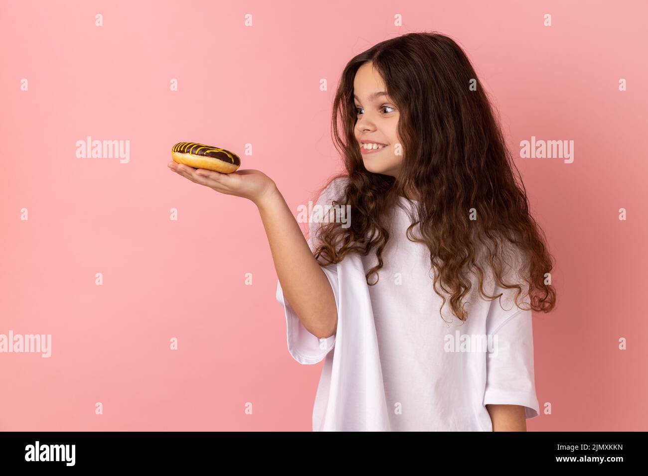 Portrait of hungry little girl wearing white T-shirt looking at donut with excited expression and want to eat confectionary, junk food. Indoor studio shot isolated on pink background. Stock Photo