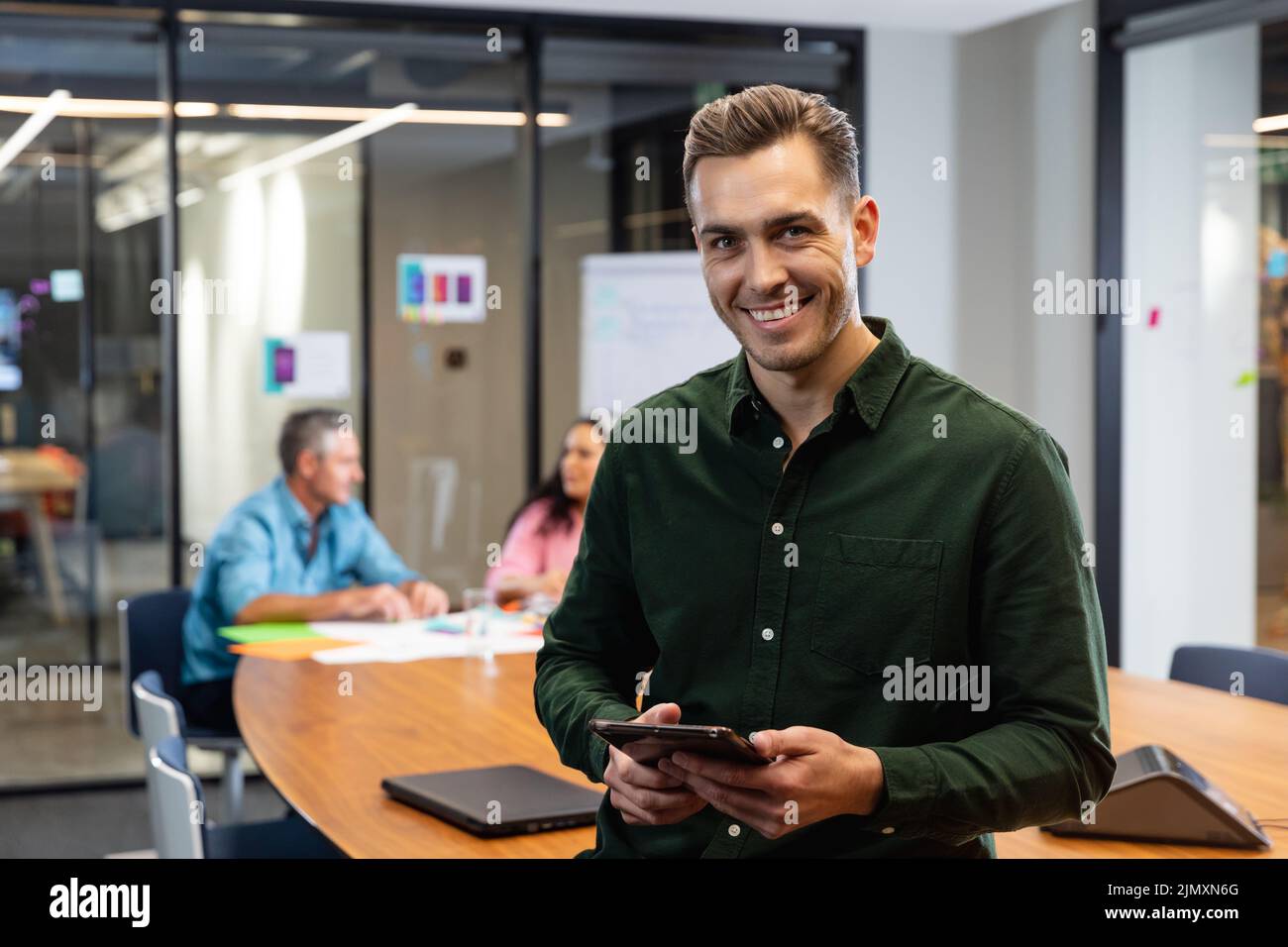 Portrait of smiling young caucasian businessman with tablet pc while colleagues in background. Unaltered, creative business, workplace, meeting, wirel Stock Photo