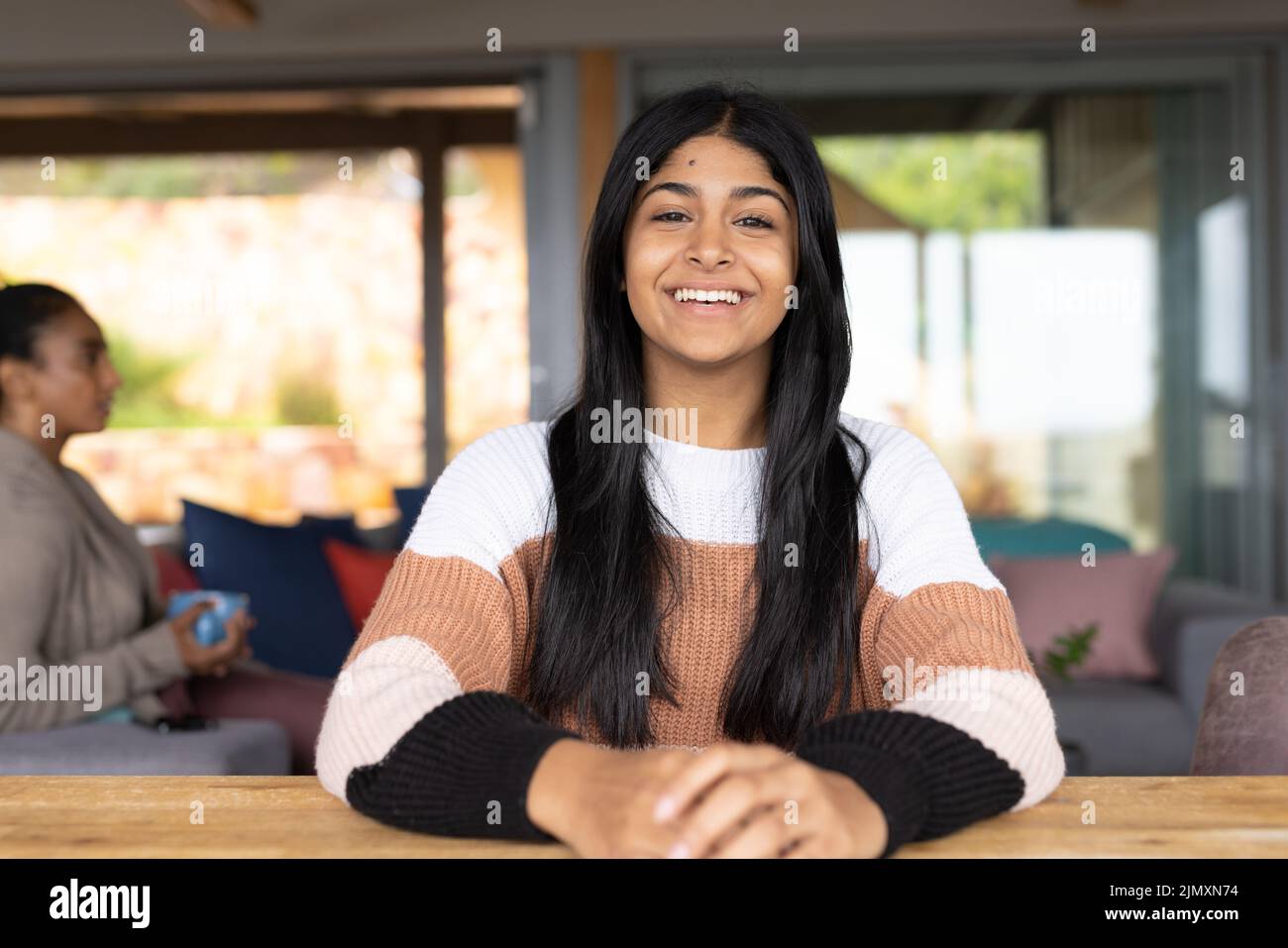 Portrait of happy biracial teenage girl sitting at table while mother with cup sitting in background Stock Photo