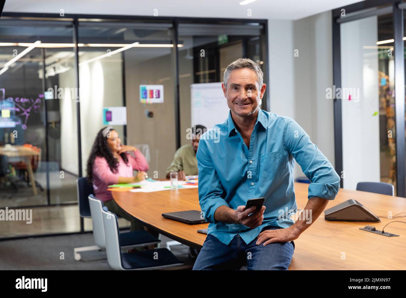 Portrait of smiling mature caucasian businessman with smart phone while colleagues in background. Unaltered, creative business, workplace, meeting, wi Stock Photo