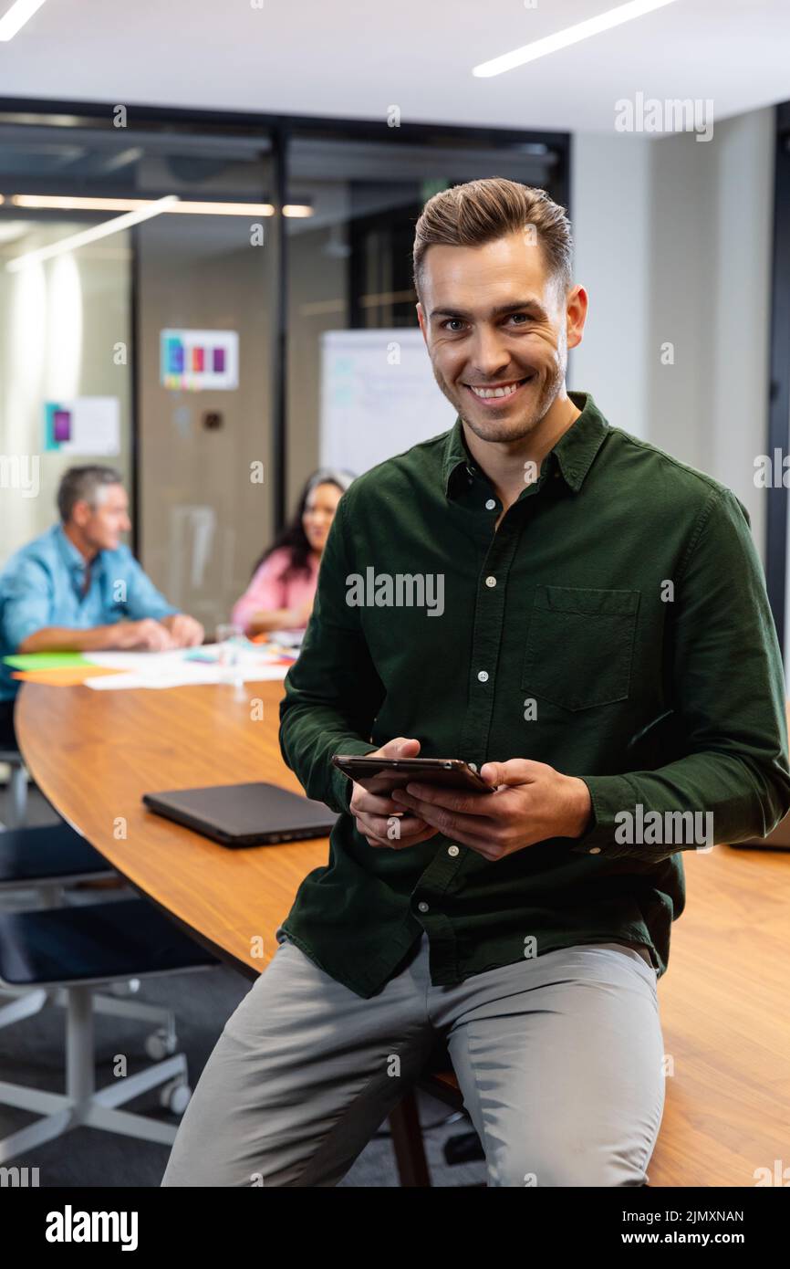 Portrait of smiling young caucasian businessman with digital tablet while colleagues in background. Unaltered, creative business, workplace, meeting, Stock Photo