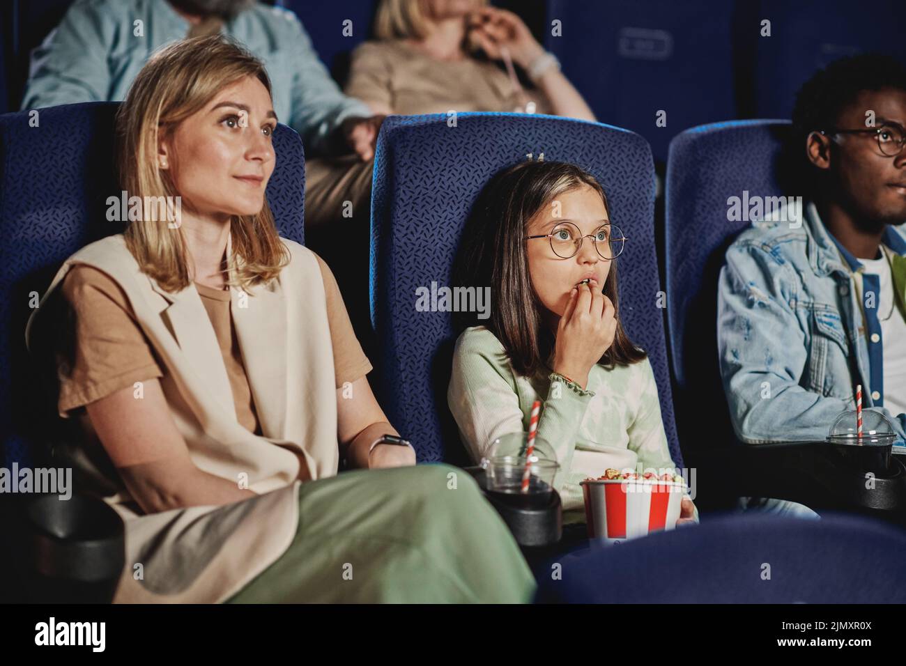 Modern Caucasian woman and her daughter spending timer together watching movie and eating popcorn at cinema Stock Photo