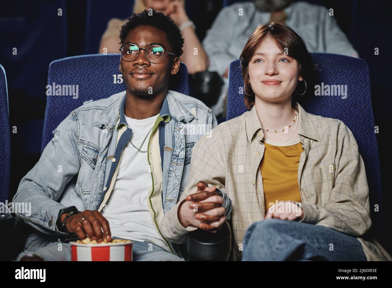 Joyful African American man and Caucasian woman in love having date at cinema watching movie and holding hands Stock Photo