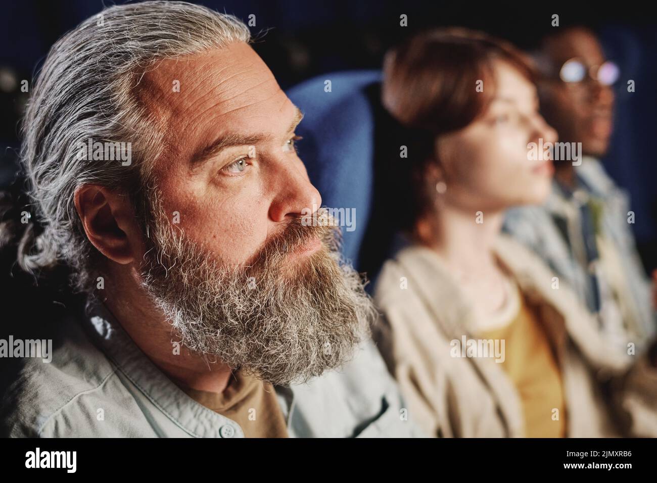 Selective focus shot of mature Caucasian man with beard on face watching drama film at cinema Stock Photo