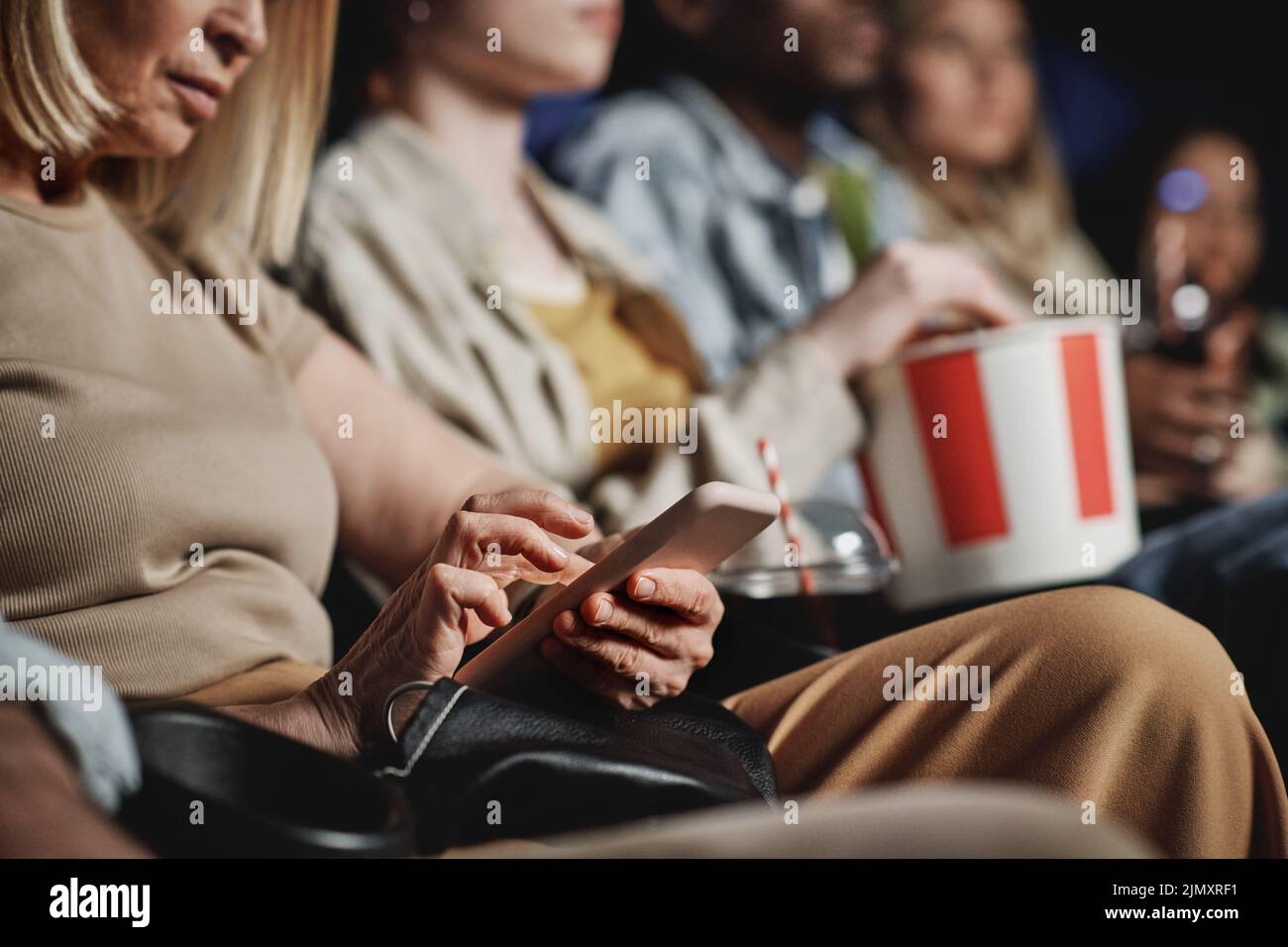 Selective focus of mature Caucasian woman surfing Internet on smartphone while watching movie at cinema Stock Photo
