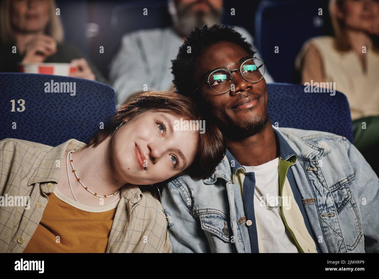 Horizontal medium close-up of lovely gen Z Black man and Caucasian woman in love leaning heads onto each other while watching movie at cinema Stock Photo
