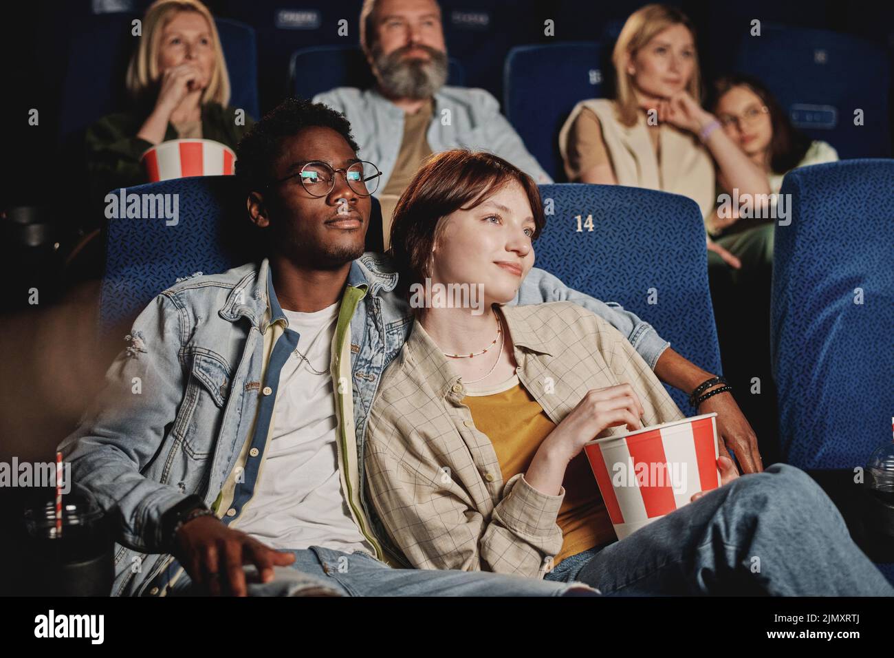 Stylish young gen Z Black man hugging his Caucasian girlfriend while watching movie together at cinema Stock Photo
