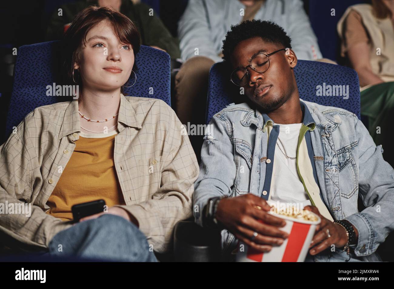 Horizontal medium portrait of young Black man having nap while his Caucasian girlfriend watching movie at cinema Stock Photo