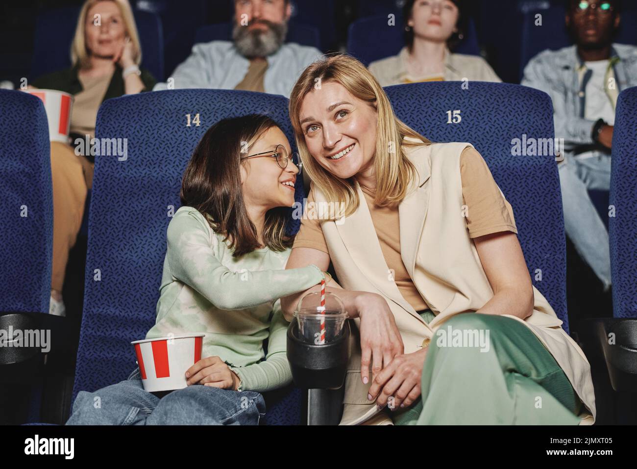 Horizontal medium shot of girl whispering something to her mother while watching move at cinema Stock Photo