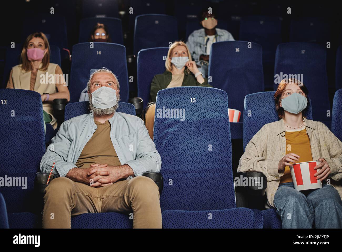 Group of people wearing masks, keeping distance from each other watching movie at cinema during quarantine Stock Photo