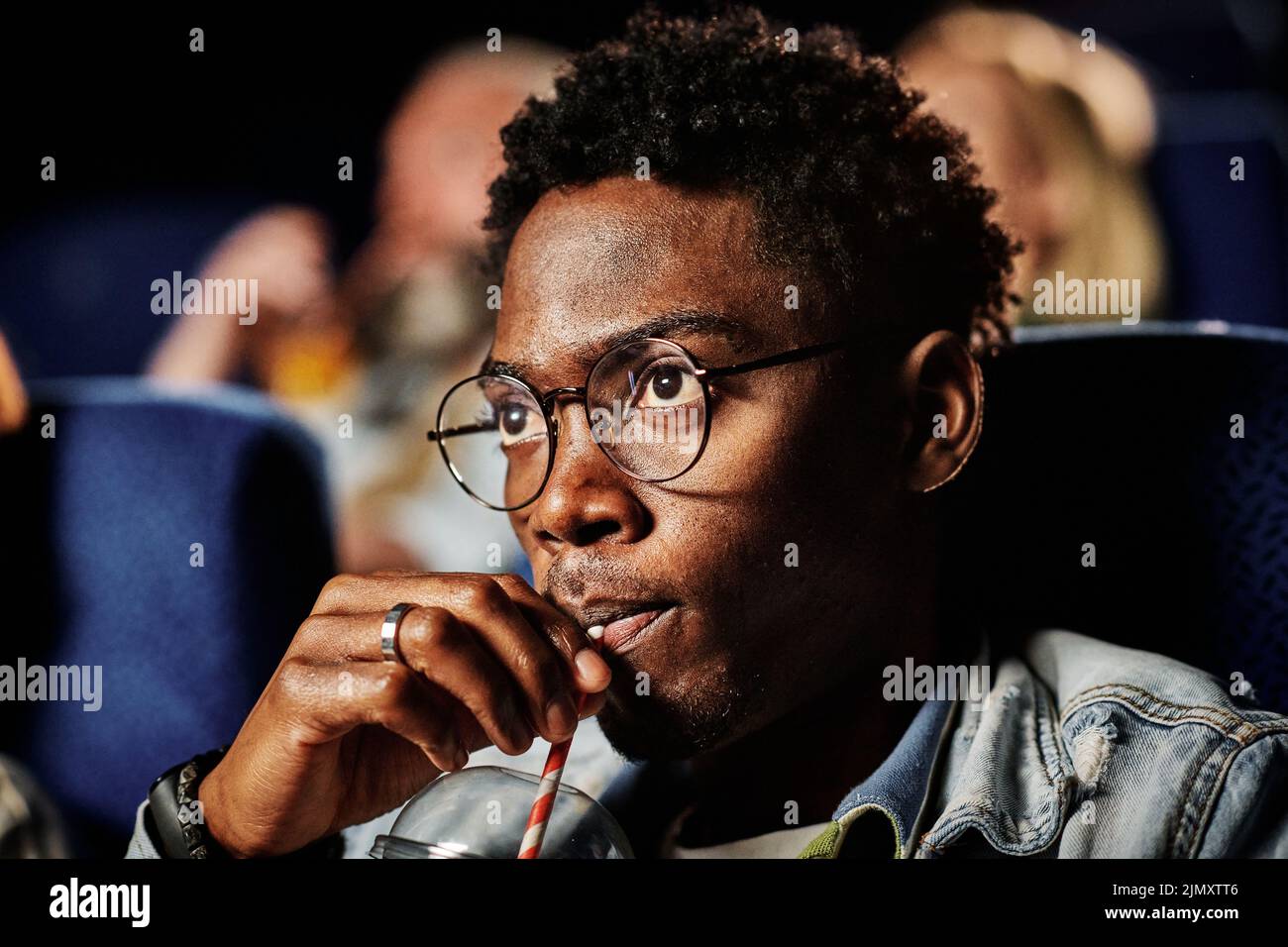 Close-up of young gen Z Black man wearing eyeglasses sipping fizzy drink while watching movie at cinema Stock Photo