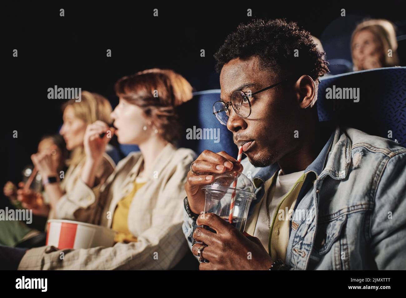 Selective focus shot of stylish young Black guy wearing eyeglasses drinking cola while watching movie at cinema Stock Photo