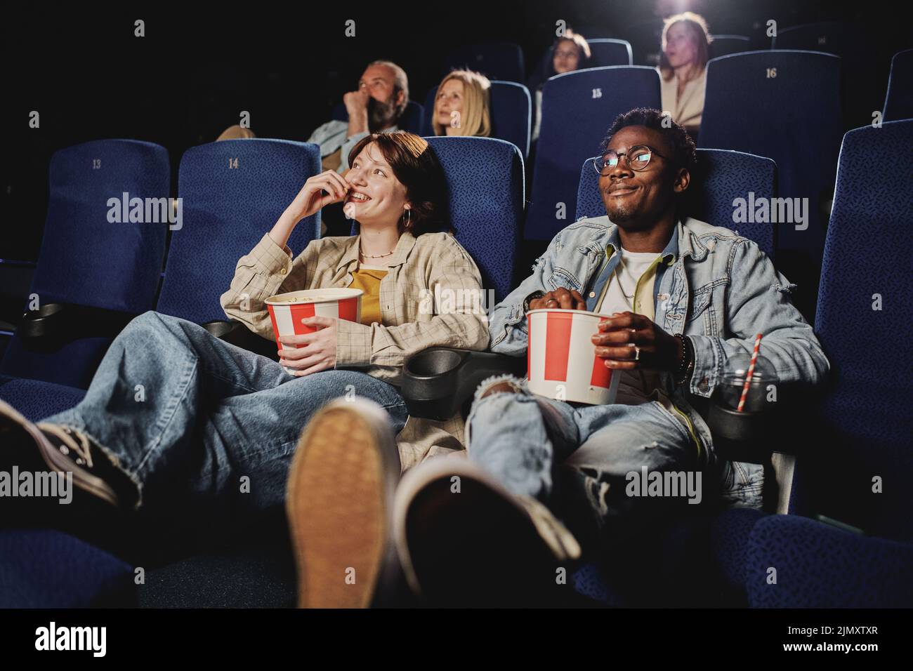 Young gen Z Black man and Caucasian woman wearing casual clothes relaxing watching movie and eating popcorn at cinema Stock Photo