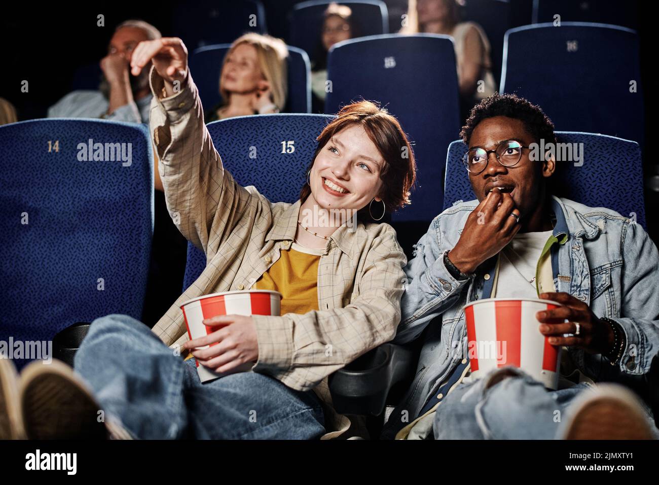 Young Caucasian woman and her Black friend spending time together at cinema watching movie and eating popcorn Stock Photo