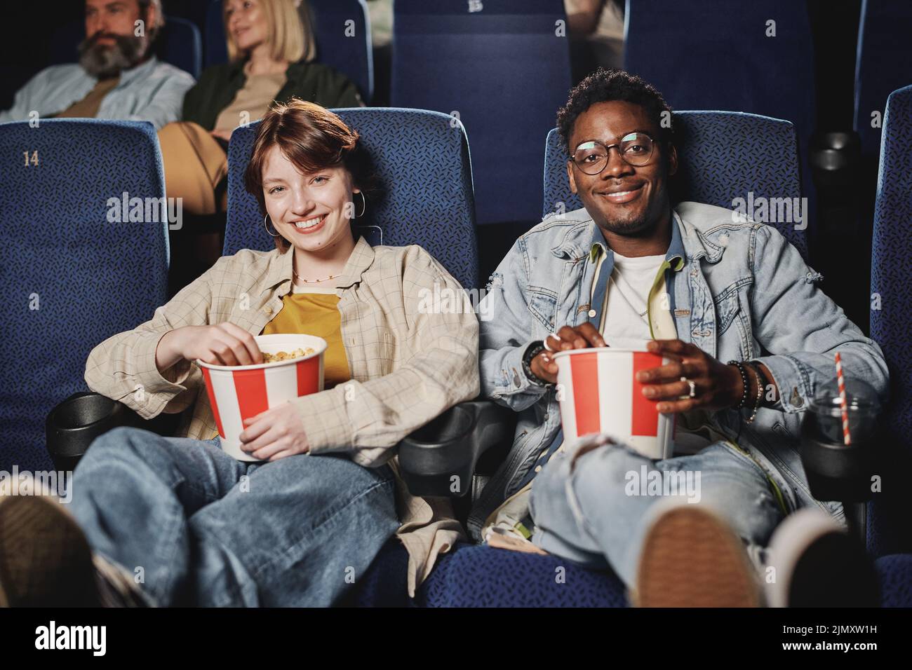 Portrait of stylish young Black man and Caucasian wiman sitting relaxed with popcorn on lap at cinema smiling at camera Stock Photo