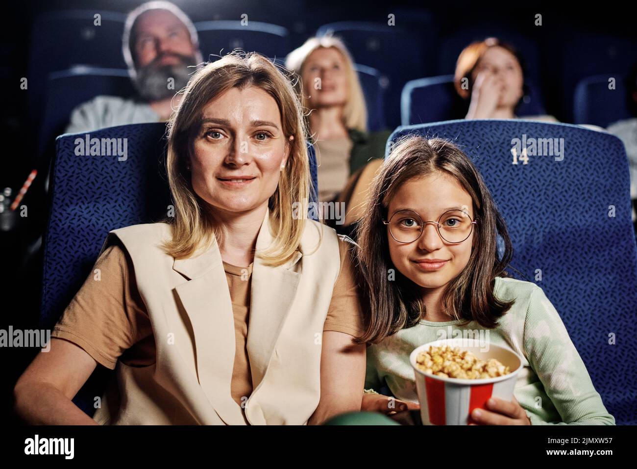 Horizontal medium portrait of joyful Caucasian mother and daughter spending time together sitting at cinema looking at camera Stock Photo