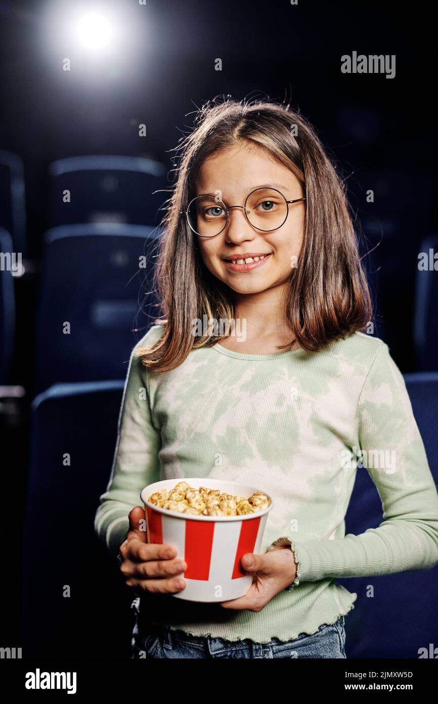 Vertical medium portrait of preteen Caucasian girl holding bucket with popcorn standing in cinema hall Stock Photo