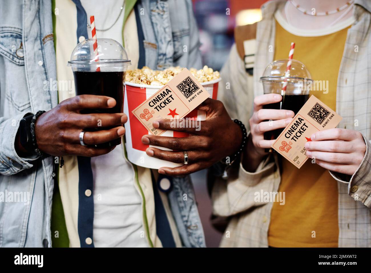 Horizontal medium section shot of unrecognizable stylish young Black man and Caucasian woman holding bucket of popcorn, cola and tickets for movie Stock Photo