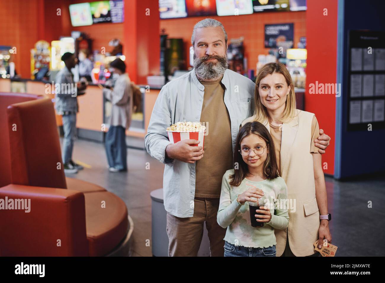 Portrait of modern family with kid holding bucket of popcorn, fizzy drink and tickets for movie looking at camera Stock Photo