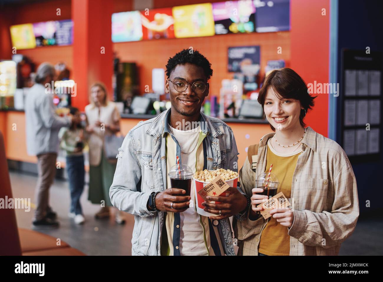 Portrait of young Black man and Caucasian woman wearing casual clothes having date at cinema holding popcorn, drinks and tickets smiling at camera Stock Photo